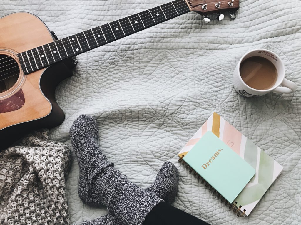flat lay of a woman in wool socks on a bed w a dreams journal, coffee and a guitar